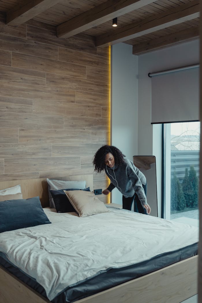 African American woman fixing pillows in a modern bedroom with wooden accents, promoting cleanliness and organization.
